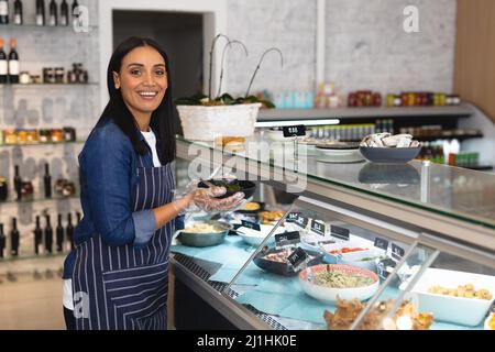 Portrait of smiling young caucasian female employee holding bowl by display cabinet in coffee shop. unaltered, cafe culture, people and occupation con Stock Photo