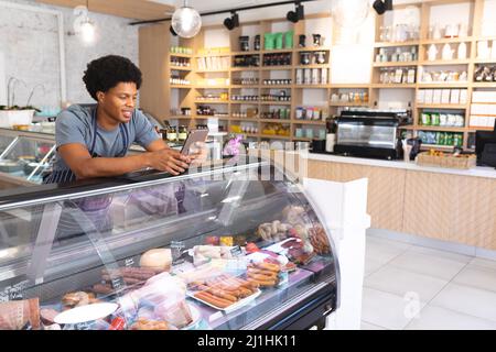 Young male african american barista using tablet pc while leaning on display cabinet in coffee shop. unaltered, cafe culture, people and occupation co Stock Photo