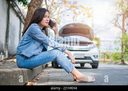 Asian woman sitting beside car after a car breakdown on street and call to car service. Concept of vehicle engine problem or accident and emergency he Stock Photo