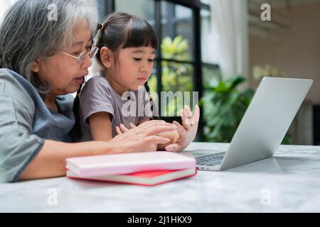 Asian Grandmother with her two grandchildren having fun and playing education games online with a computer notebook at home in the living room. Concep Stock Photo