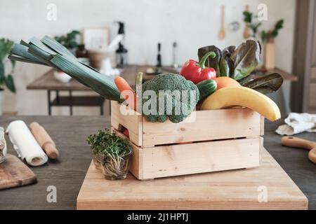 Wooden box full of various vegetables and fruits placed on board with growing plants into glasses in modern kitchen Stock Photo