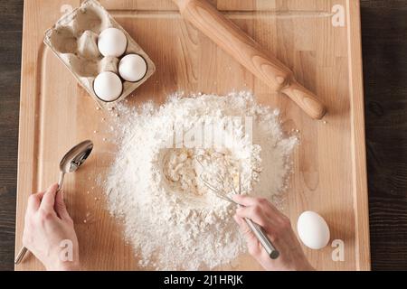 Above view of unrecognizable woman mixing flour and egg with whisk on wooden board with rolling pin Stock Photo