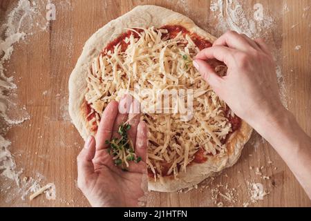 Above view of unrecognizable woman adding herbs on raw pizza placed on wooden surface with flour Stock Photo