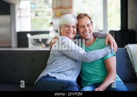 I love having him offer. Portrait of a senior woman embracing her son while sitting on the sofa at home. Stock Photo