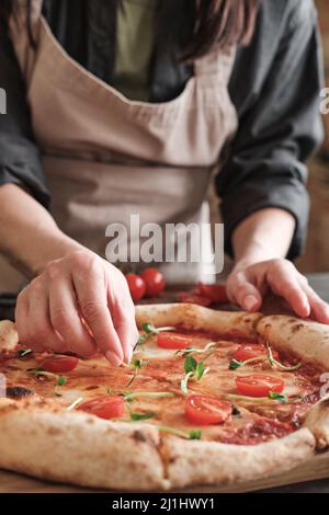 Close-up of unrecognizable woman in apron putting pea leaves on pizza with tomatoes while preparing it for family Stock Photo