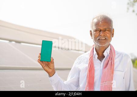 smiling farmer showing green screen mobile phone by pointing finger while standing in front of solar panel at farmland - concept of app advertisement Stock Photo
