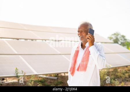 happy Indian farmer talking on mobile phone in front of solar panels about electric power generation farmland - concept of technology and modern Stock Photo