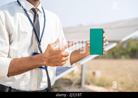 hands showing green screen mobile phone by pointing finger in front of solar panel - concept of application promotion, booking service and Stock Photo