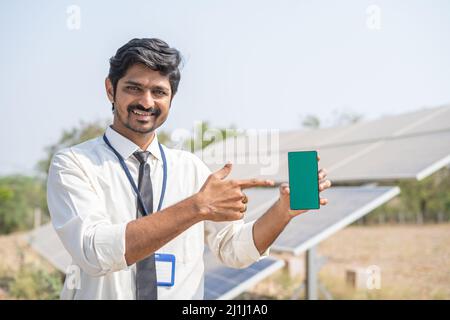 happy engineering standing in front of solar panel showing green screen mobile phone by looking camera at farmland - concept of application promotion Stock Photo