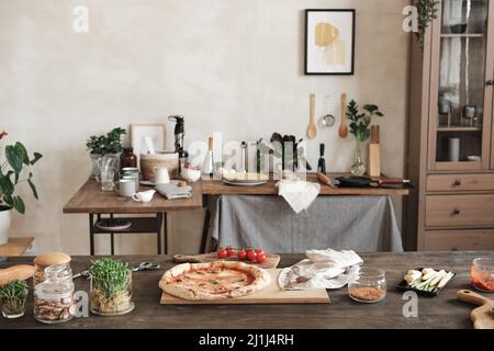 Prepared pizza on wooden board placed on table with ingredients and jars of spices, table with utensils and crockery in background Stock Photo