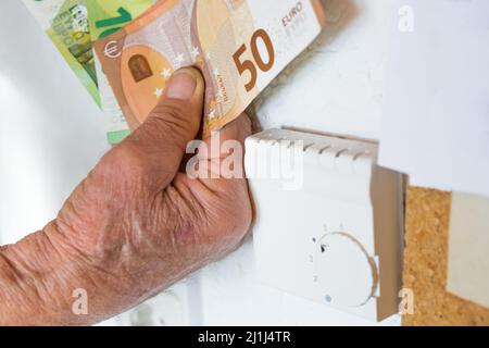 Old pensioner man holding banknotes in hand in front of a heating thermostat in an apartment, Germany Stock Photo
