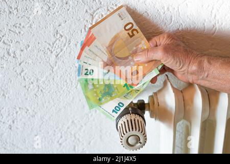 Old senior citizen man holding banknotes in hand in front of a heating thermostat and radiator in an apartment, Germany Stock Photo
