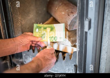 Old pensioner man holding banknotes in hand in front of wood stove with wood and burning lighter in an apartment, Germany Stock Photo