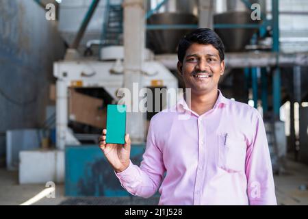 supervisor showing green screen mobile phone while looking at camera at factory infront of machinery - concept of booking service, promotion and Stock Photo
