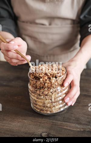 Close-up of unrecognizable woman in apron standing at wooden table and preparing granola with muesli Stock Photo