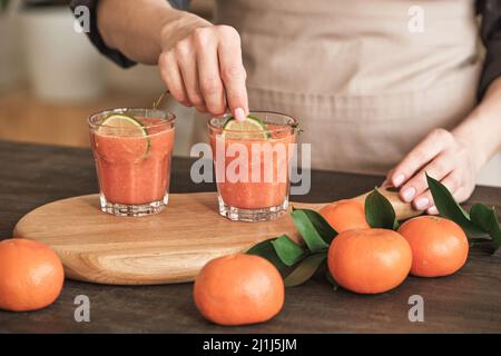 Close-up of unrecognizable woman in apron standing at table and adding lime slices into tangerine cocktails on wooden board Stock Photo