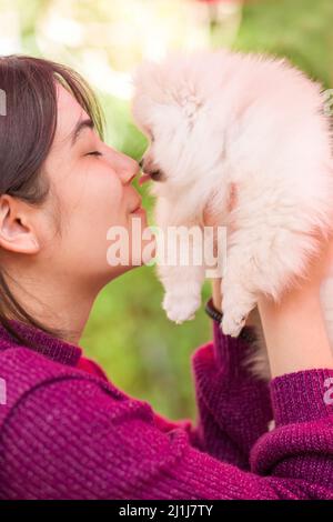 Biracial young woman in purple sweater holding small white pomeranian puppy outdoors on deck Stock Photo