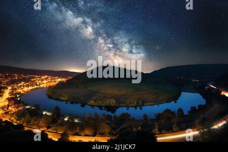 Majestic night sky with the Milky Way over a river bend around a hill, framed with an illuminated road and town Stock Photo