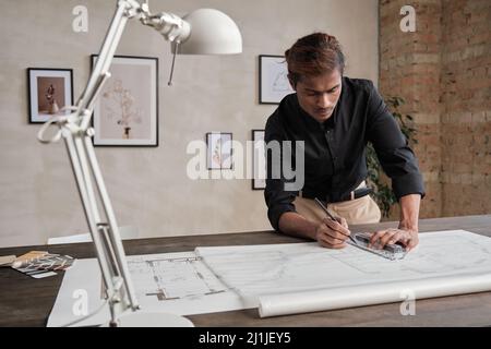 Serious young Black architect in shirt standing at wooden desk and drawing with ruler while working on new project Stock Photo