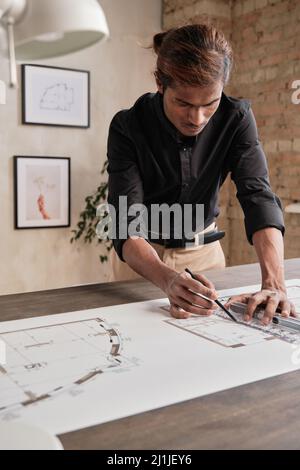 Busy young Indian man in black shirt standing at table and using ruler while drafting house plan in loft office with minimalistic pictures on wall Stock Photo