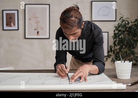 Busy young mixed race engineer in black shirt standing at table and leaning on it while using ruler and drawing line on floor plan Stock Photo