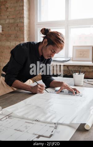 Focused young mixed race engineer with ponytail leaning on desk and using ruler while drawing house plan in office Stock Photo