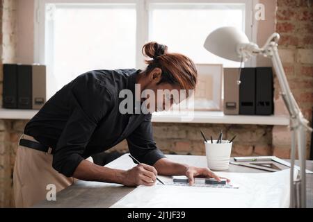 Serious middle-aged Black designer with red hair standing at wooden desk and editing sketch with pencil while working on blueprint Stock Photo