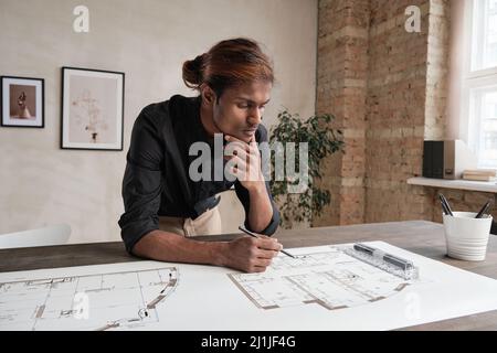 Thoughtful young mixed race architect with dyed hair leaning on table and analyzing sketch while thinking about design Stock Photo