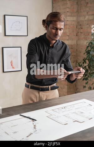 Serious young Indian architect in black shirt standing at table and photographing blueprint of house on smartphone Stock Photo
