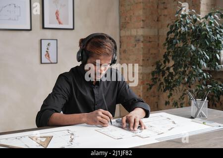 Concentrated young Indian architect in wireless headphones sitting at table and listening to music while working on blueprint Stock Photo