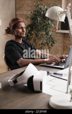 Content young Indian architect with ponytail sitting at wooden desk with virtual reality headset and developing design on laptop in loft office Stock Photo