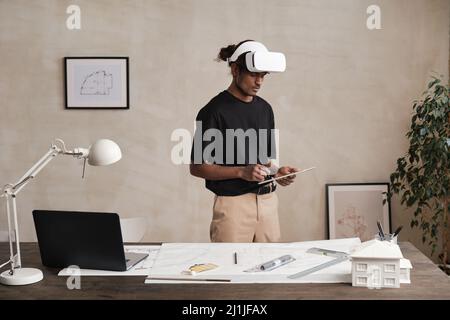 Serious busy young Indian architect in virtual reality headset standing in modern office and analyzing online sketch Stock Photo