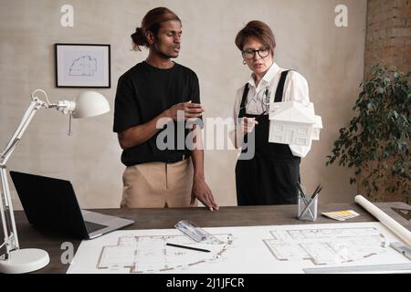 Serious young Indian architect with ponytail gesturing hand while presenting private house project to lady boss holding D model of house Stock Photo