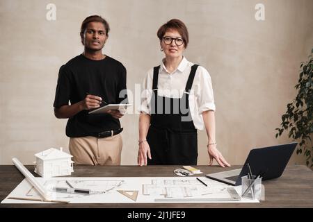Portrait of young Indian architect with tablet and female engineer in bodysuit standing at table with supplies and blueprint Stock Photo