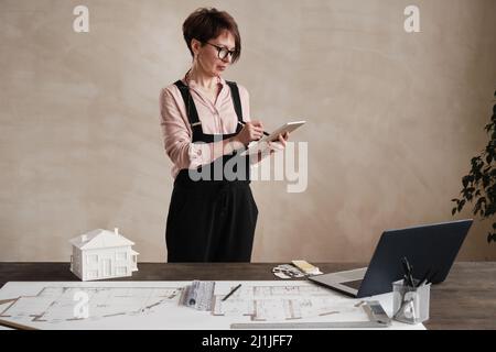 Content mature architect with short hair standing at desk with laptop, D model of house and blueprint and drawing on tablet Stock Photo