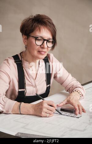 Positive mature Caucasian architectural designer in eyeglasses sitting at table and drawing line using ruler Stock Photo