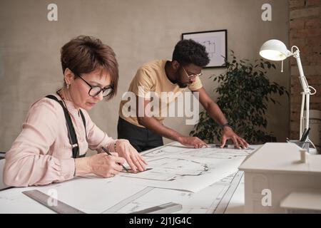 Concentrated mature woman in glasses sitting at table and creating drawing for architectural project together with African-American intern Stock Photo