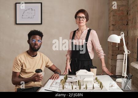 Portrait of multi-ethnic architects in eyewear drinking coffee and working with housing prototype in loft office Stock Photo