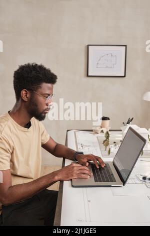 Serious young African-American building manager with beard sitting at desk and using laptop while working with online data Stock Photo