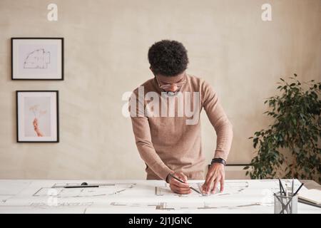 Serious young African-American architect in glasses standing at desk and drafting blueprint while working on building project Stock Photo