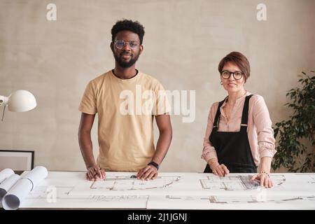 Team of positive interracial architects standing at desk with floor plan and rolls of blueprints in modern office with minimalistic interior Stock Photo