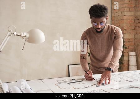 Serious young Black man in brown turtleneck and glasses standing at table and using ruler and pencil while preparing technical drawing Stock Photo
