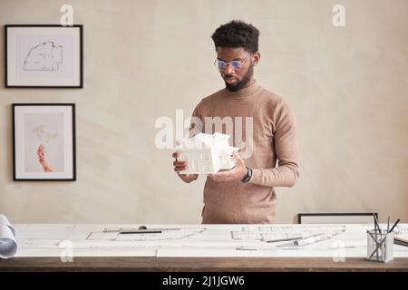Serious young black architect with beard standing at table with floor plan and analyzing D model of house in office Stock Photo