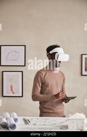 Modern young African-American architect in VR goggles standing in office with blueprints and D model on table and using digitizer Stock Photo