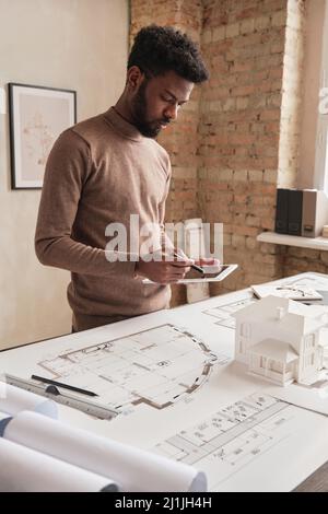 Serious young African-American architectural engineer in brown sweater standing at table with blueprint and examining design project plan on tablet Stock Photo