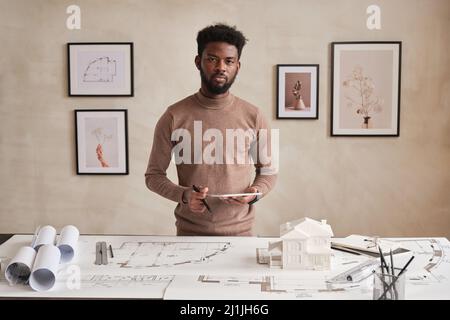 Portrait of serious young bearded black architect in sweater standing with tablet at table full of sketches and supplies in contemporary office Stock Photo