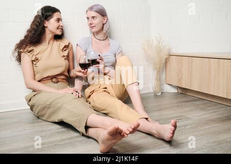 Happy young Caucasian lesbians sitting on floor and clinking wineglasses while drinking wine at home Stock Photo