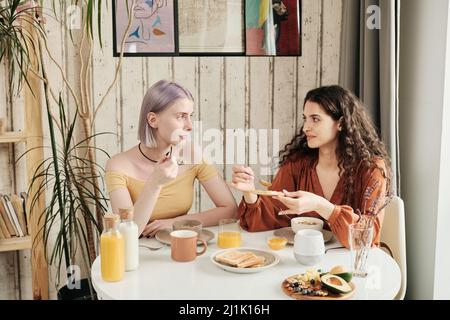 Happy young lesbians sitting at dining table and chatting while eating toasts with jam at home Stock Photo