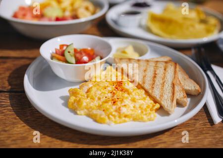 Varied breakfast on wooden table. Close-up view on scrambled eggs, toast bread and vegetable salad. Stock Photo