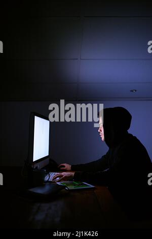 Scouring the net for his next victim. Shot of a hooded computer hacker using a computer in the dark. Stock Photo